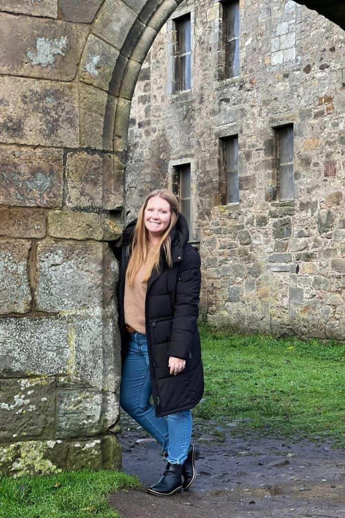Me in Scotland at Midhope Castle Tayllor, a Europe travel blogger,in a black coat leaning against a stone castle wall at Midhope Castle., one of my favorite places in Europe- Scotland!