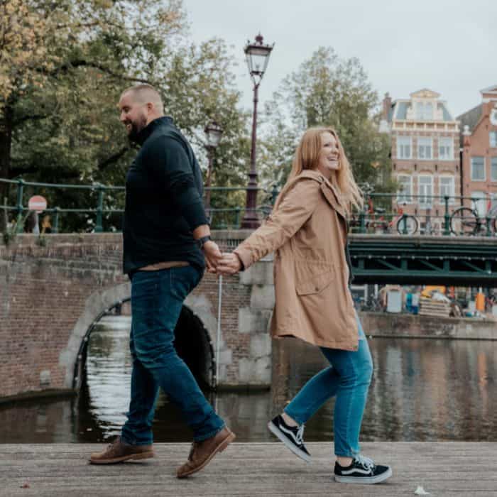 Taylor, who helps you travel Europe easily, holding hands with her husband and jumping apart. in Amsterdam. A bridge is behind them and a canal bridge.
