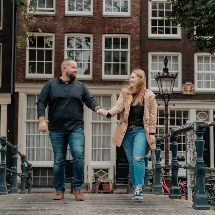 Amsterdam Travel Taylor and her husband holding hands and walking along a bridge in Amsterdam, one of the most featured places on this Europe travel blog. Red and brown canal houses are visible in the background.